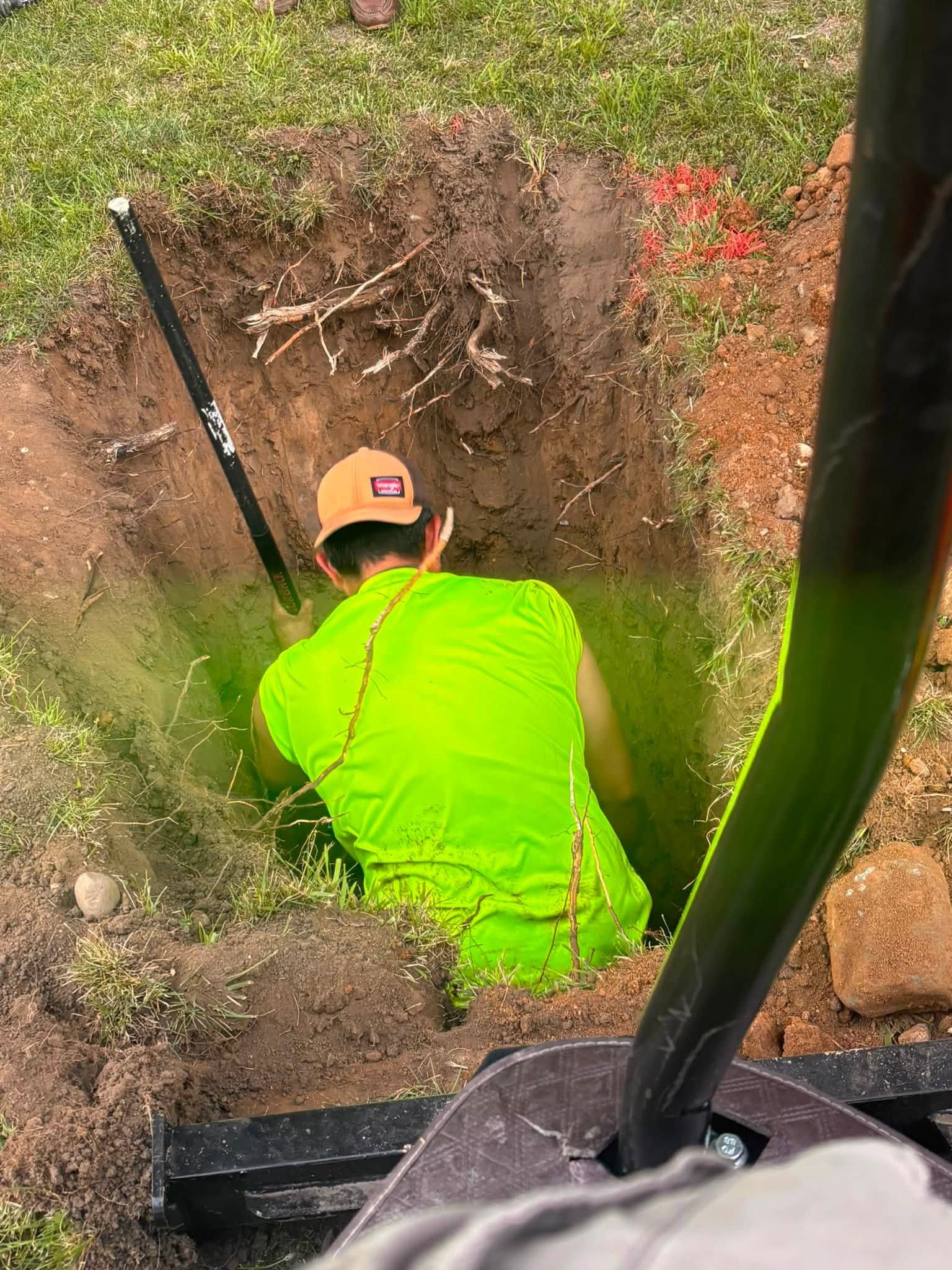 Worker in neon green shirt in a narrow dirt trench beside a yard, digging near exposed roots.