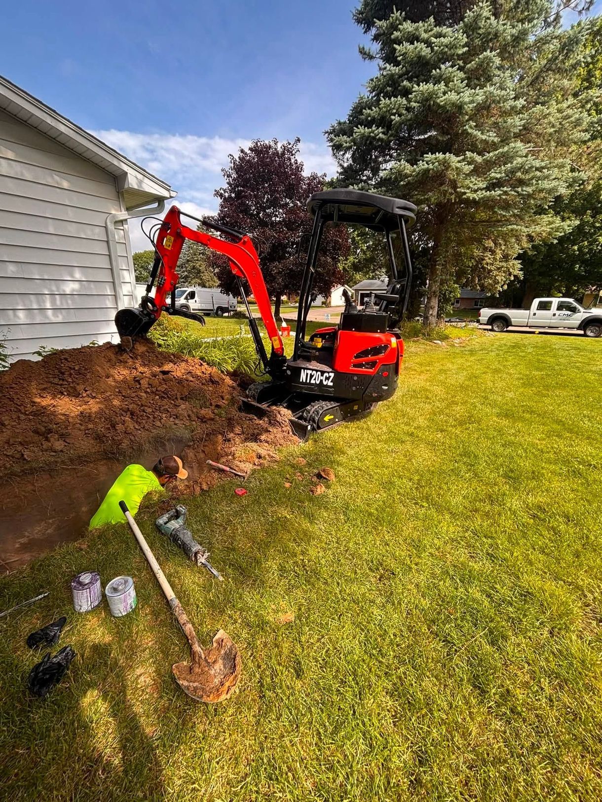 Red mini excavator by a house spreading mulch along the foundation in a sunny yard.