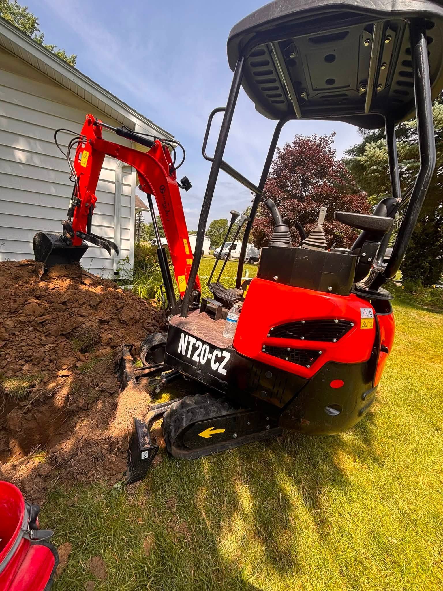 Mini excavator digging beside a house, with a red arm and black cab on grassy ground.