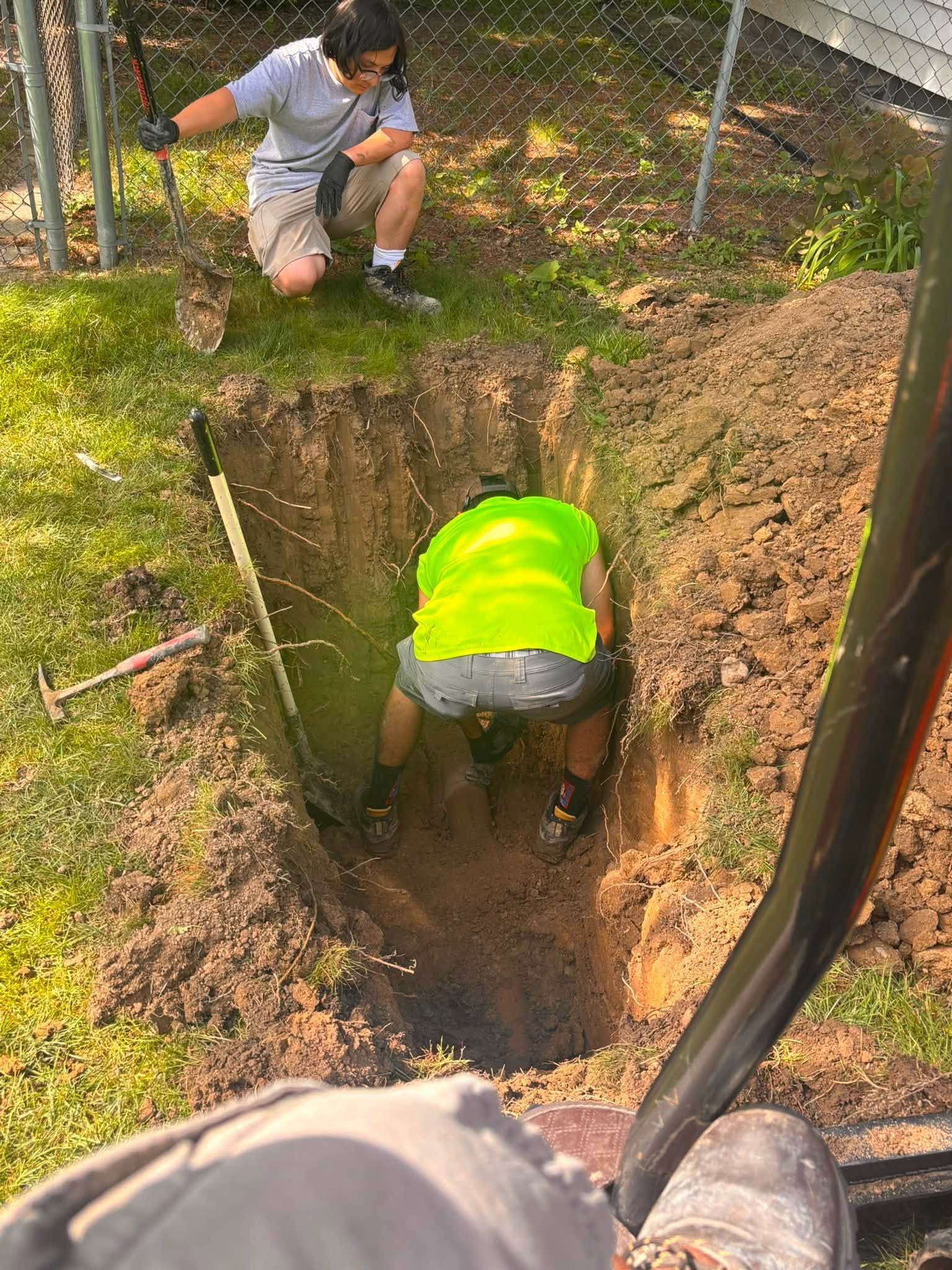 Two workers inspect a deep narrow trench in a fenced dirt area outdoors.