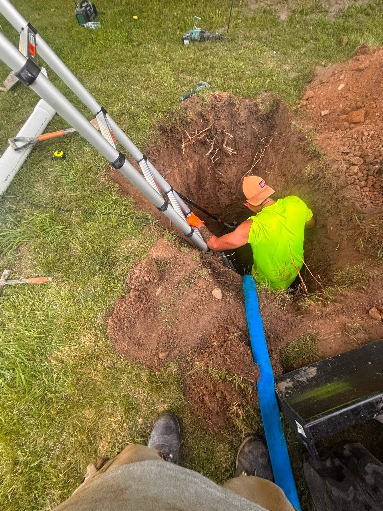 Worker in a bright neon shirt in a dirt trench, handling a blue hose beside a ladder.