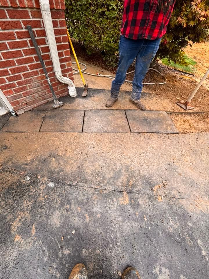 Person standing beside brick wall and sidewalk with a yellow broom and a shovel near debris.