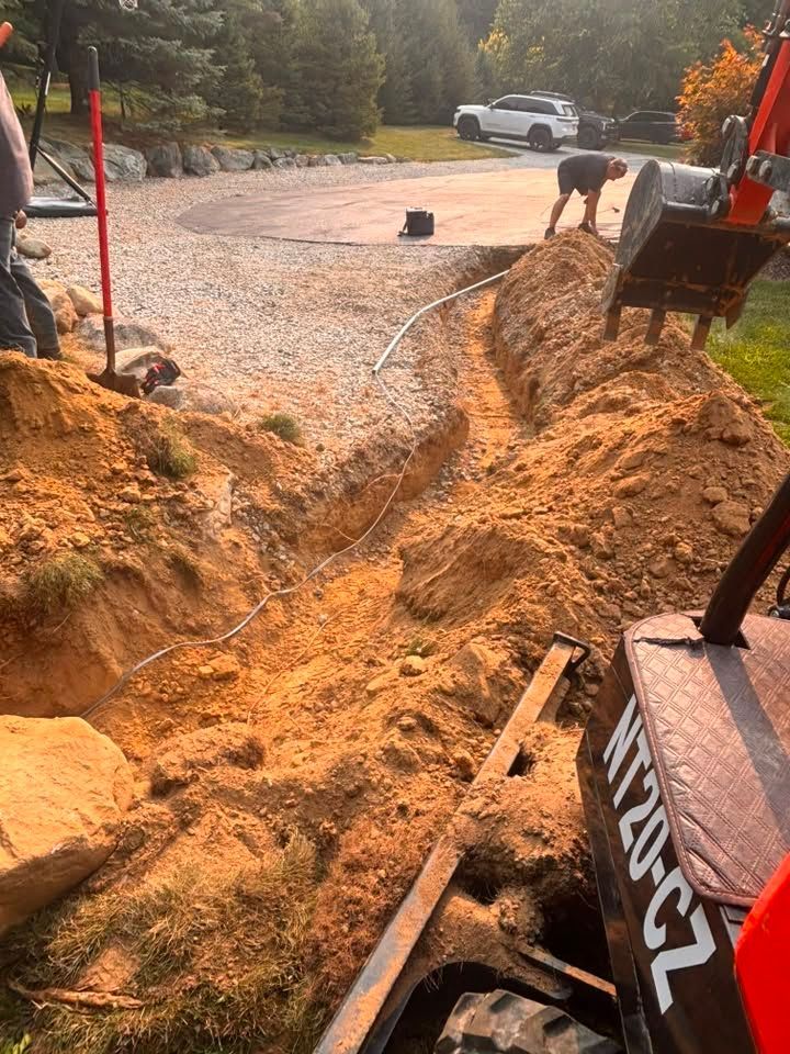 Construction trench beside a gravel road, with an excavator, hose, and worker near a white pickup truck.
