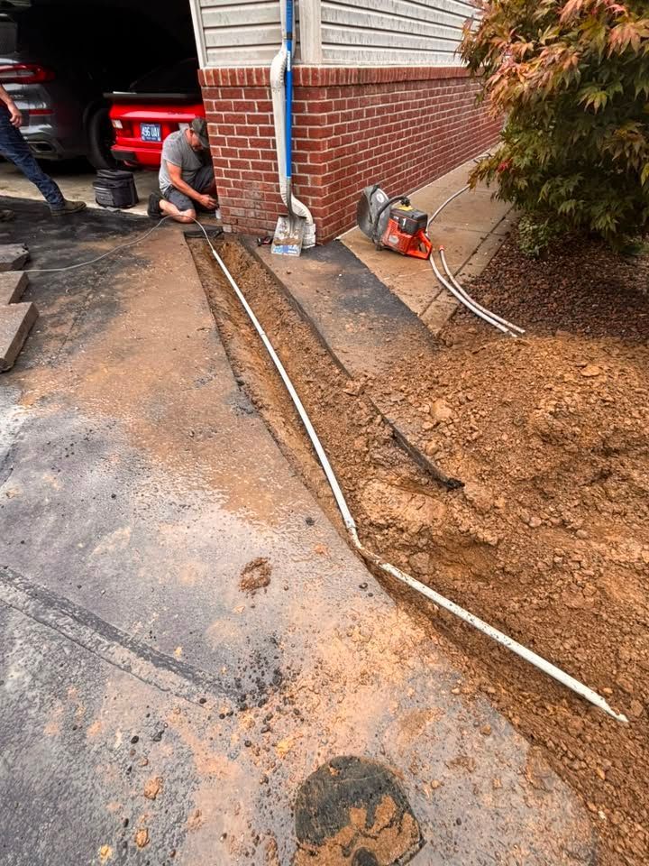 Worker operating a concrete saw beside a brick house, cutting a trench in muddy driveway after rain