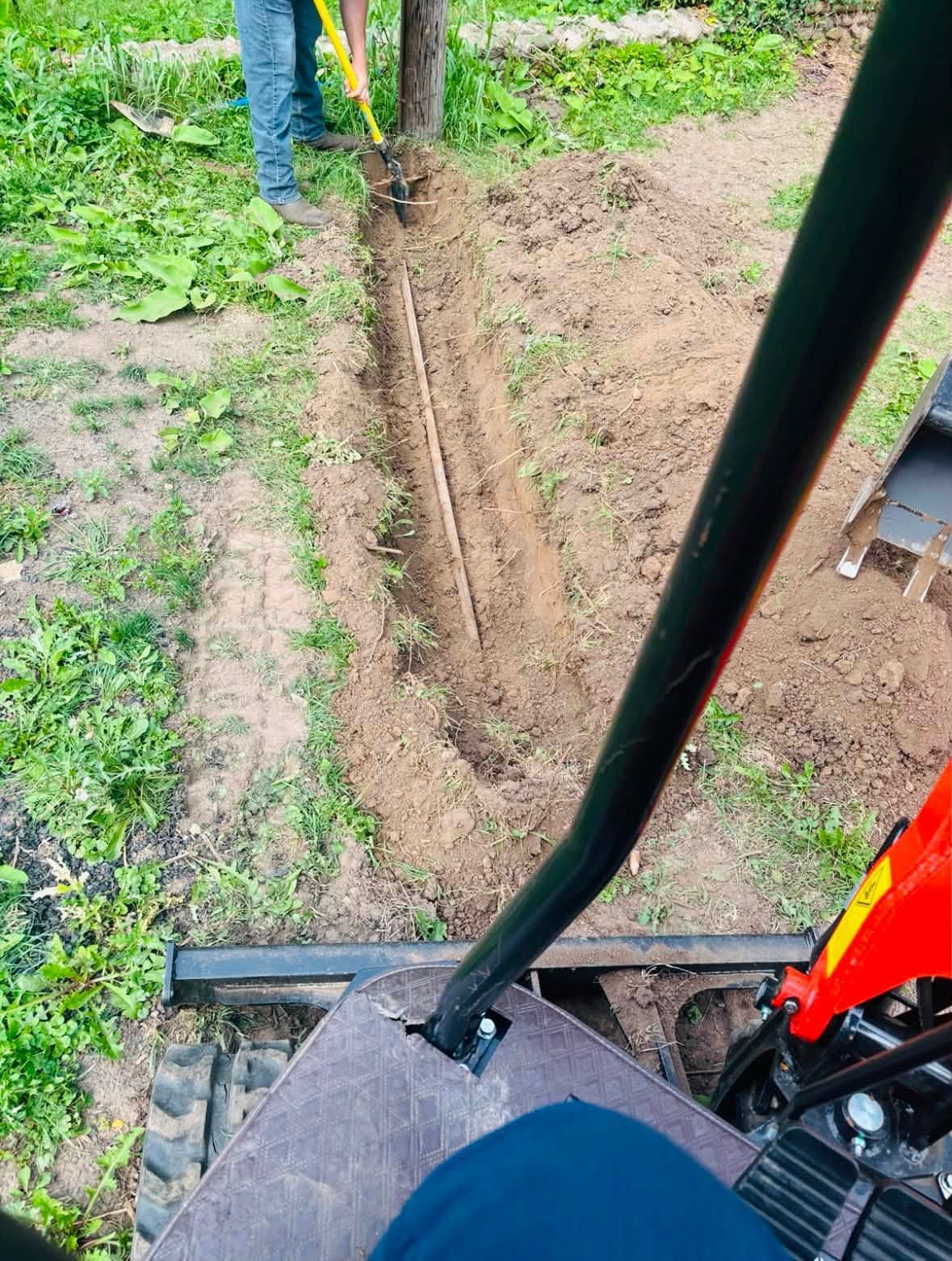 Excavator arm digging a trench beside grass, with a worker standing nearby
