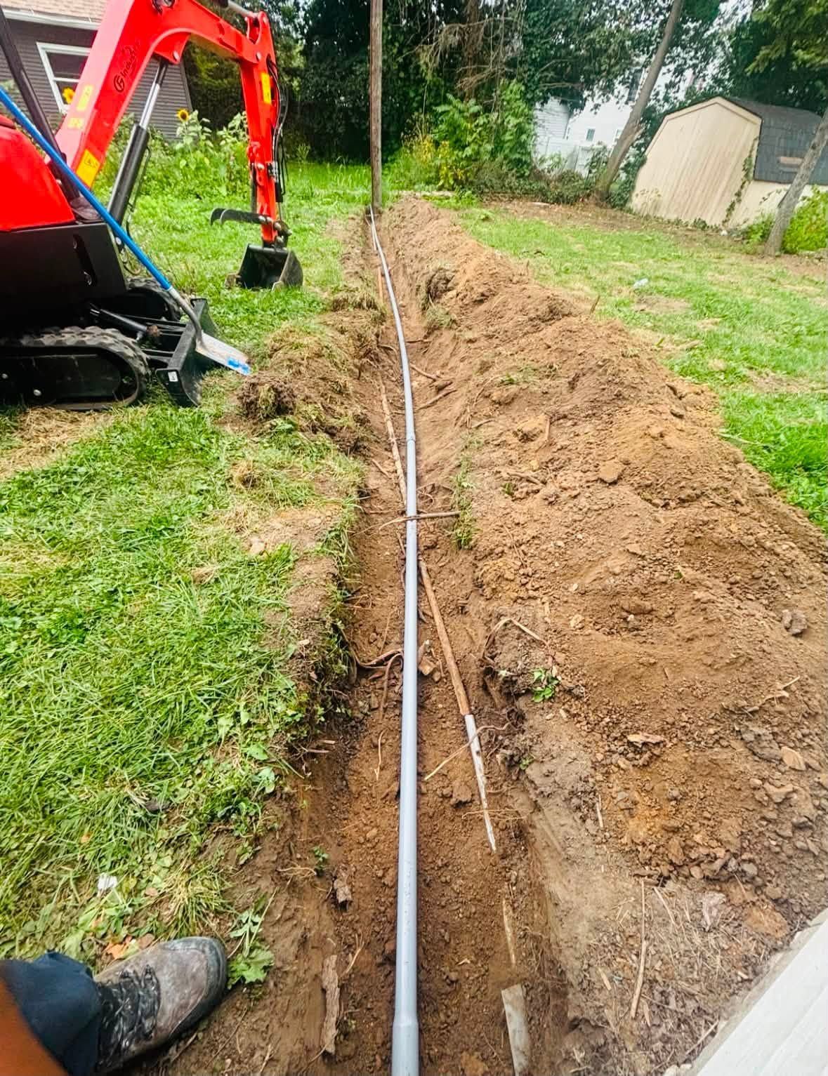 Backhoe trenching a grassy yard beside a long white pipe or cable
