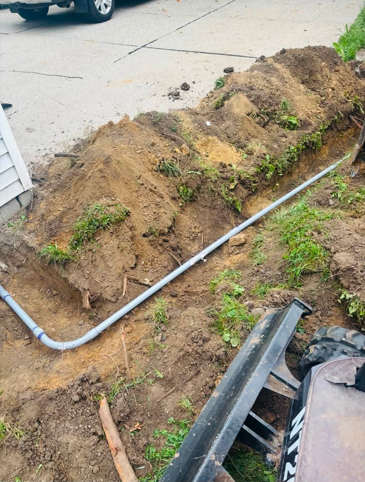 Broken metal pipe exposed in dirt beside a driveway and foundation wall.