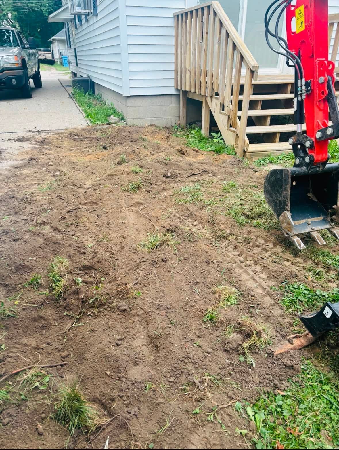 Bare dirt yard beside a house, with a red excavator bucket near wooden steps.