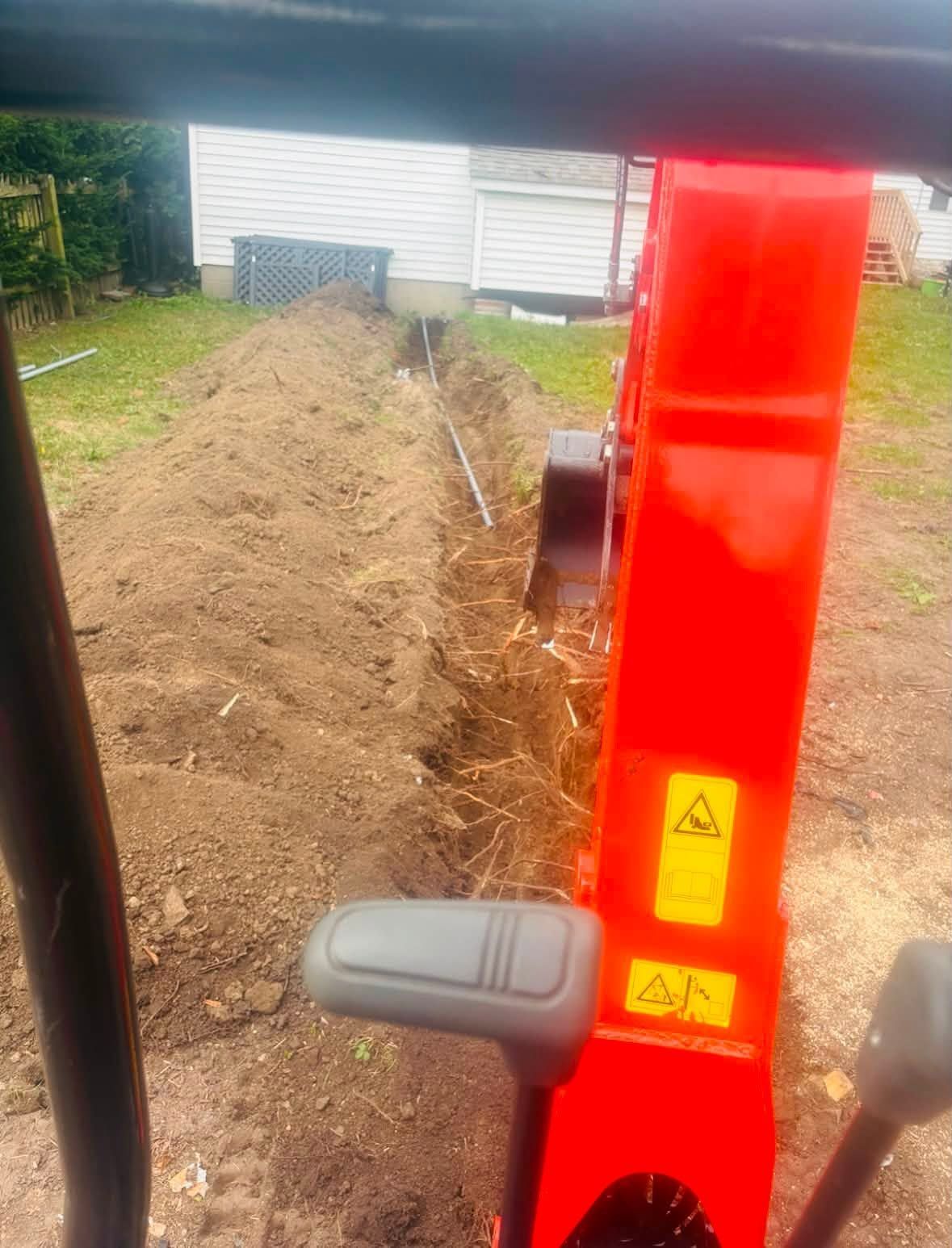Red excavator arm beside a dirt trench near a white building and grassy yard.