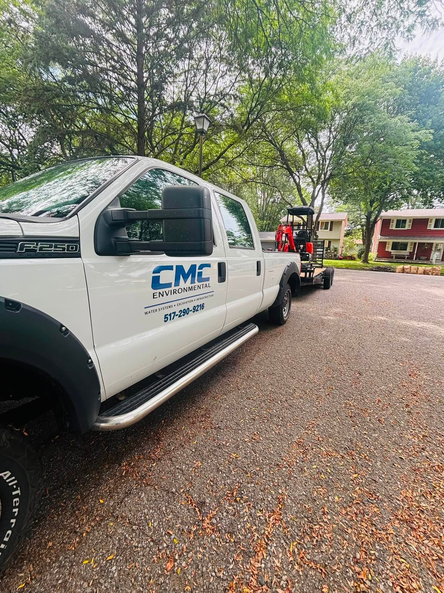 White CME truck parked on a gravel driveway under trees, with a person in a red shirt near a trailer.