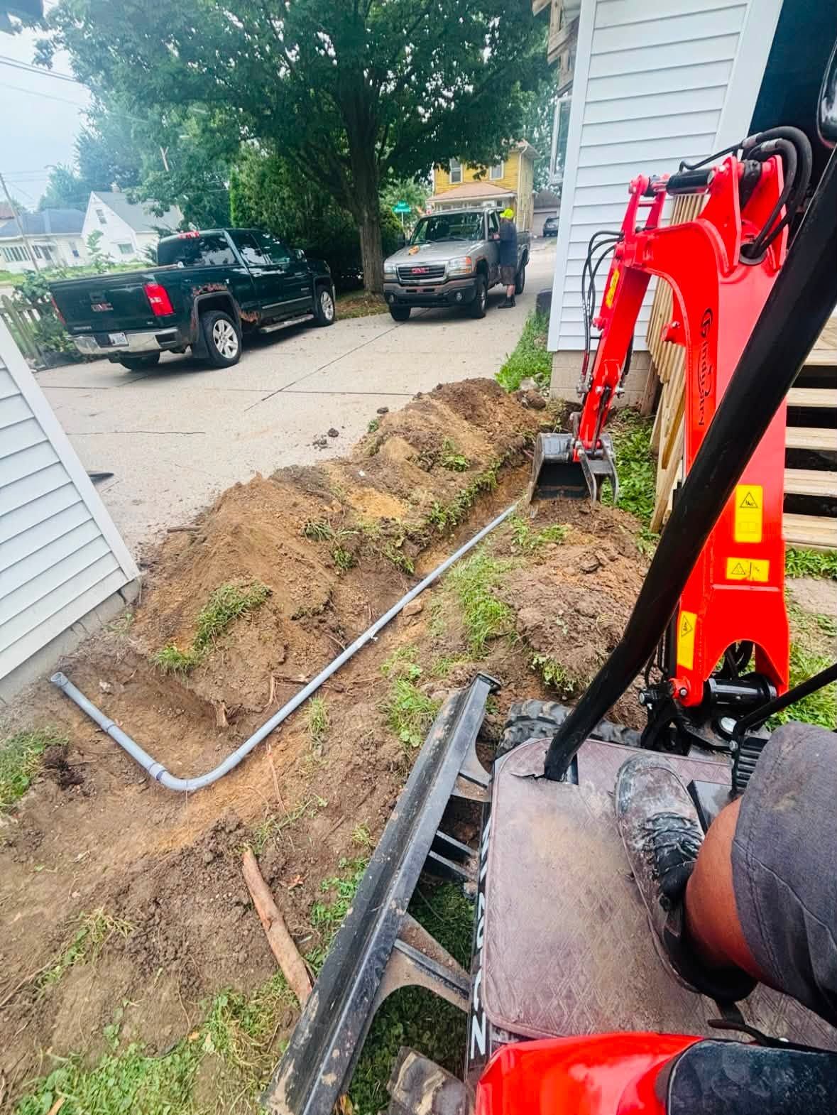 Excavator digging a trench along a house beside a driveway with parked trucks and exposed pipe.