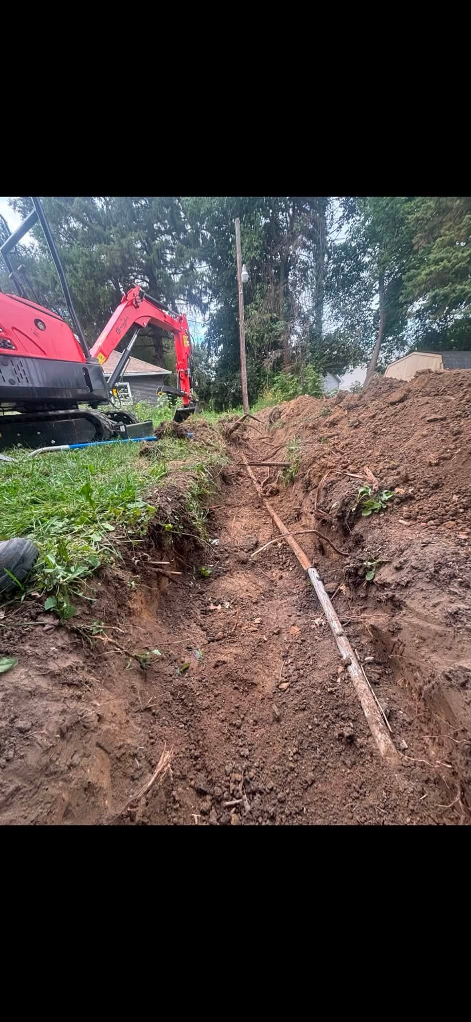 Red excavator digging a narrow trench beside a dirt mound in a wooded area.