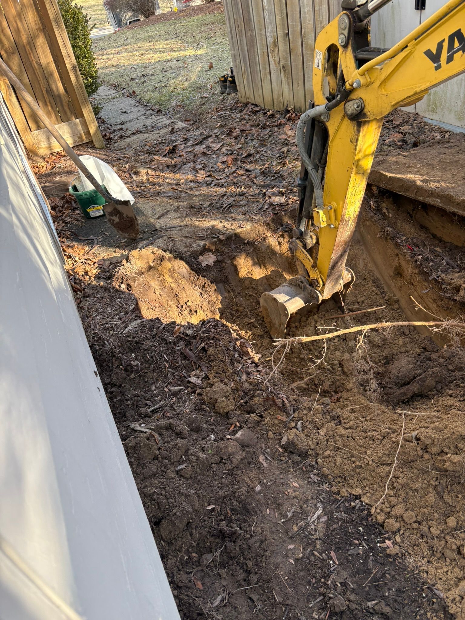 Yellow excavator digging a trench beside a fence, with dirt piled around the worksite