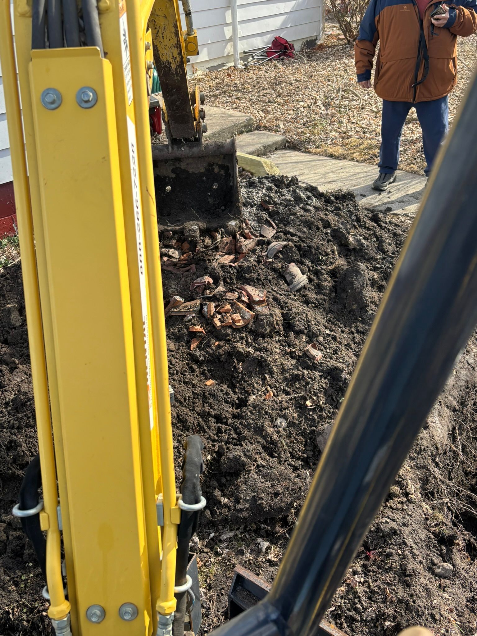 Yellow excavator arm digging up soil near a worker in an orange jacket.