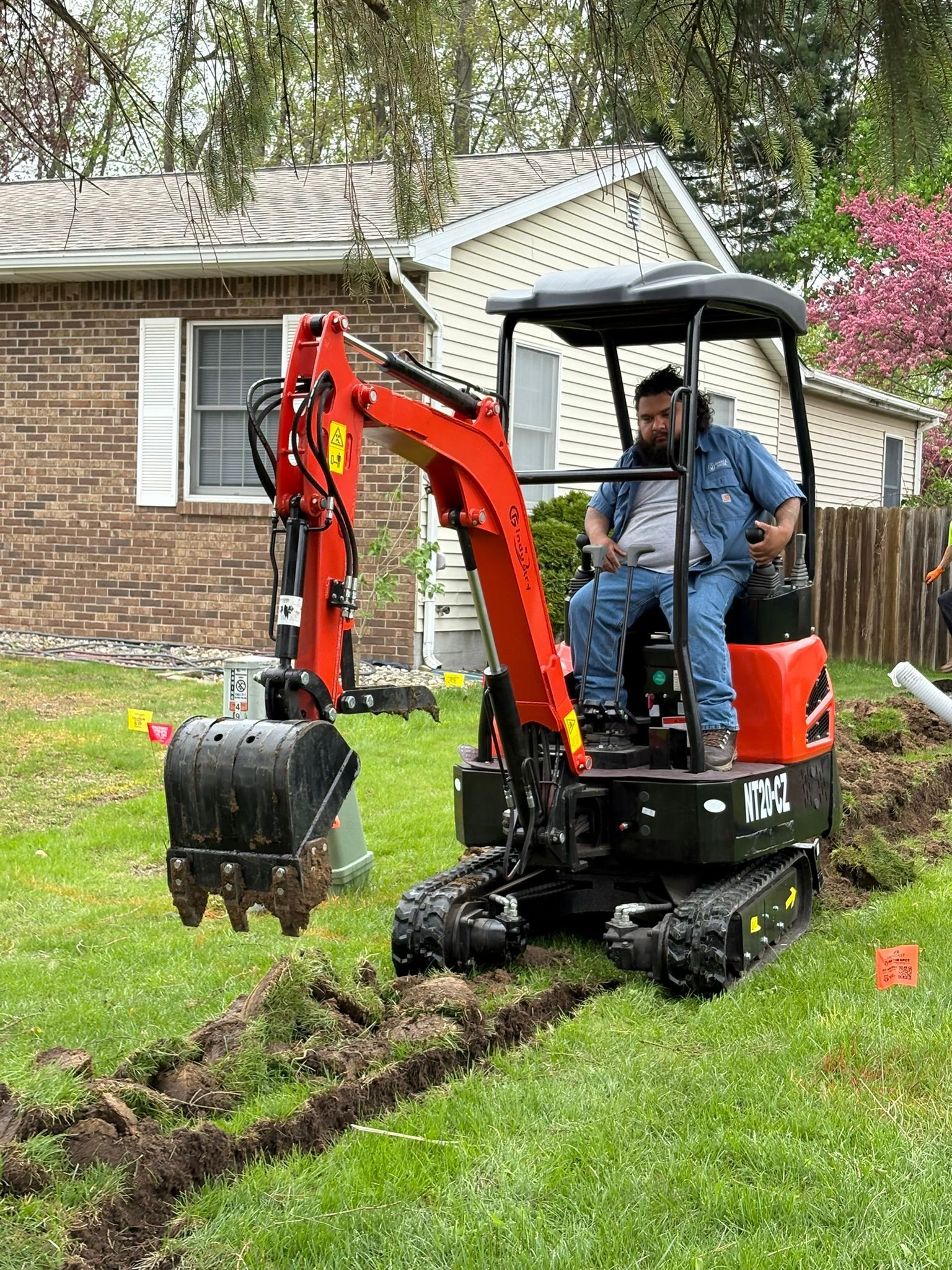 Mini excavator digging a trench in a grassy yard beside a brick house