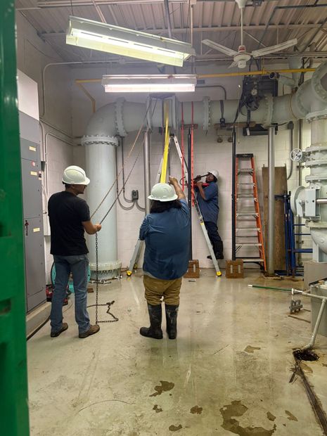 Workers in hard hats inspect a wet construction site with ladders, tools, and exposed ceiling fixtures.