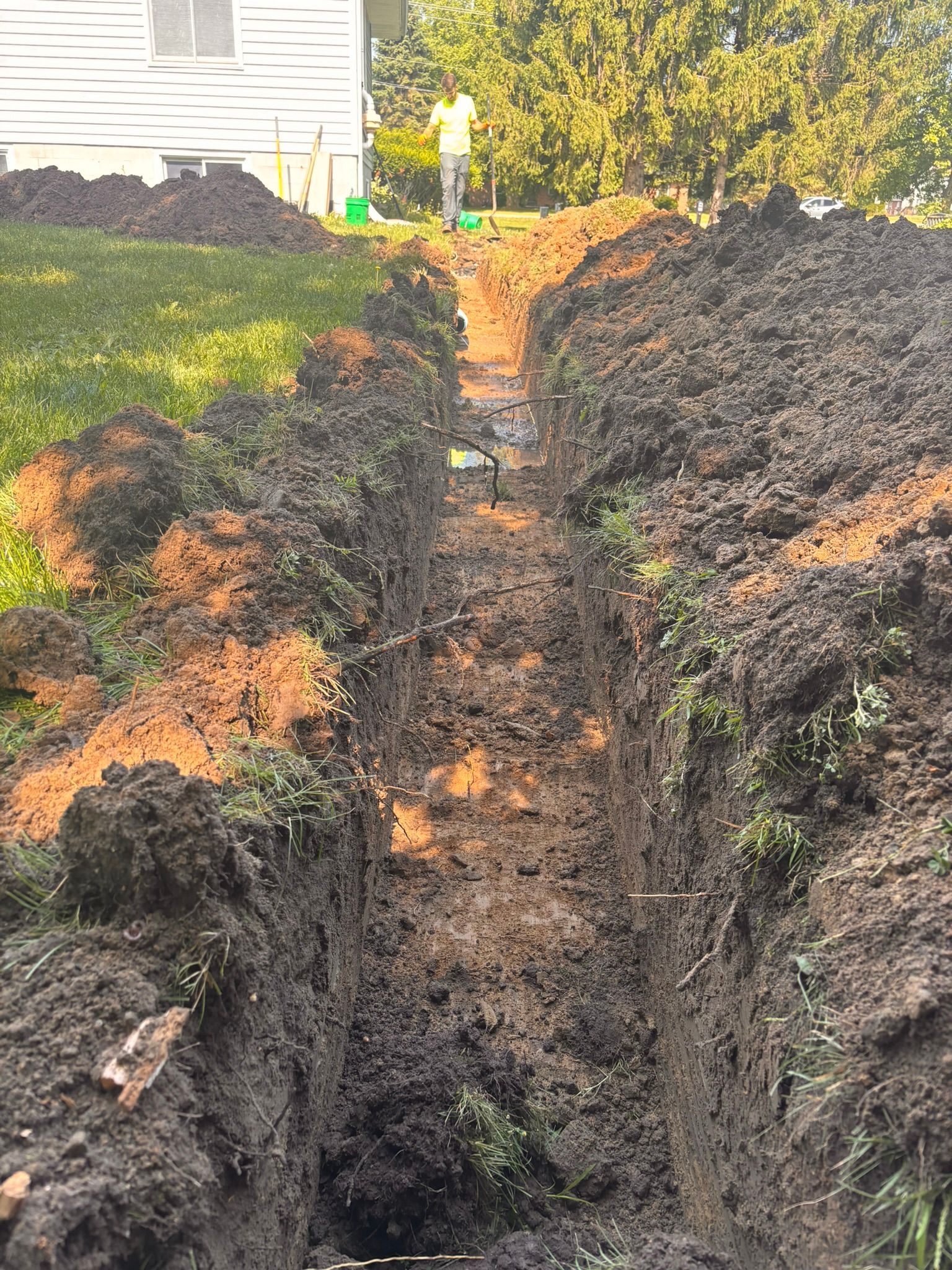 Freshly dug trench through a yard beside a house, with piles of dark soil and a narrow path of dirt.