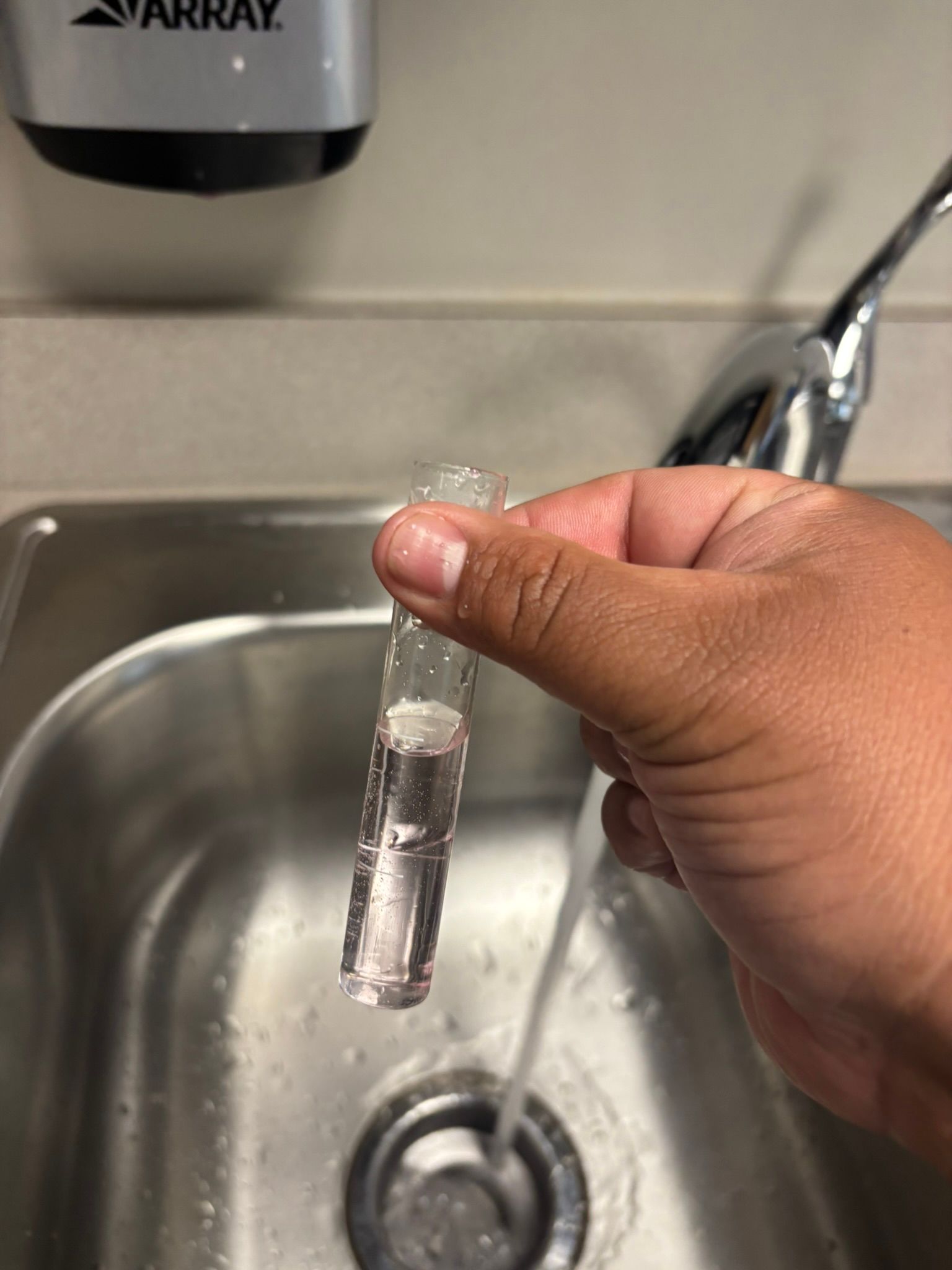 Hand holding a clear plastic test tube under a running sink faucet.
