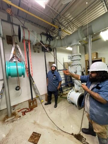 Two workers inspecting industrial piping and equipment in a mechanical room, one pointing upward.