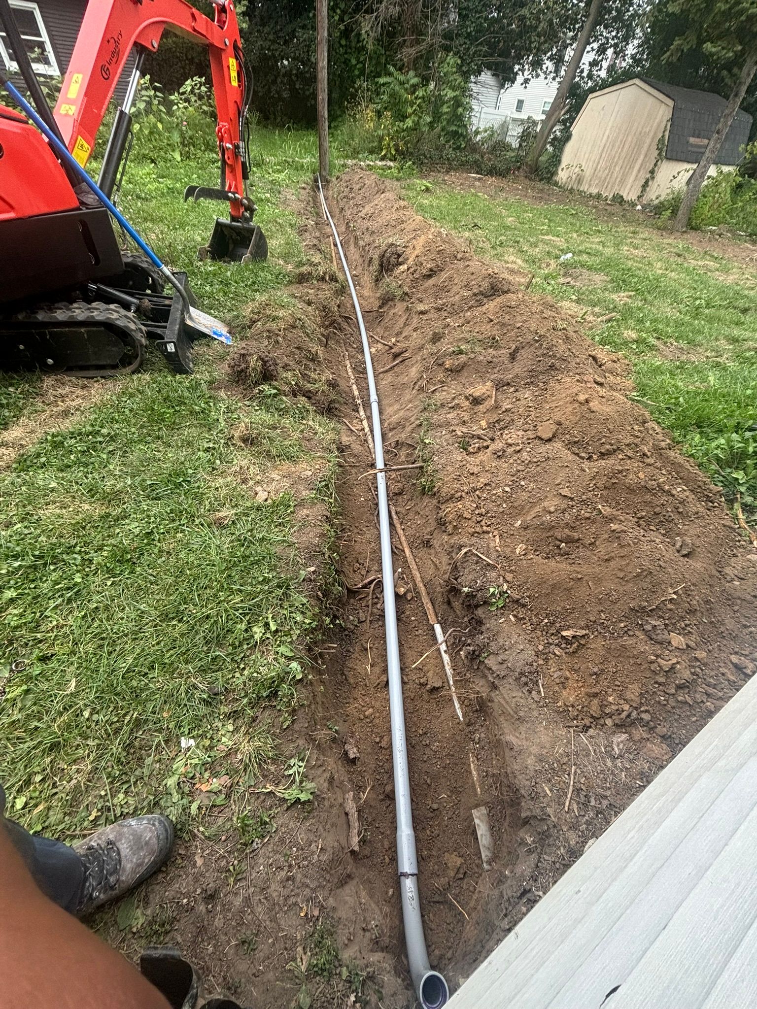 Backyard trench with a long pipe laid beside an excavator and fresh soil