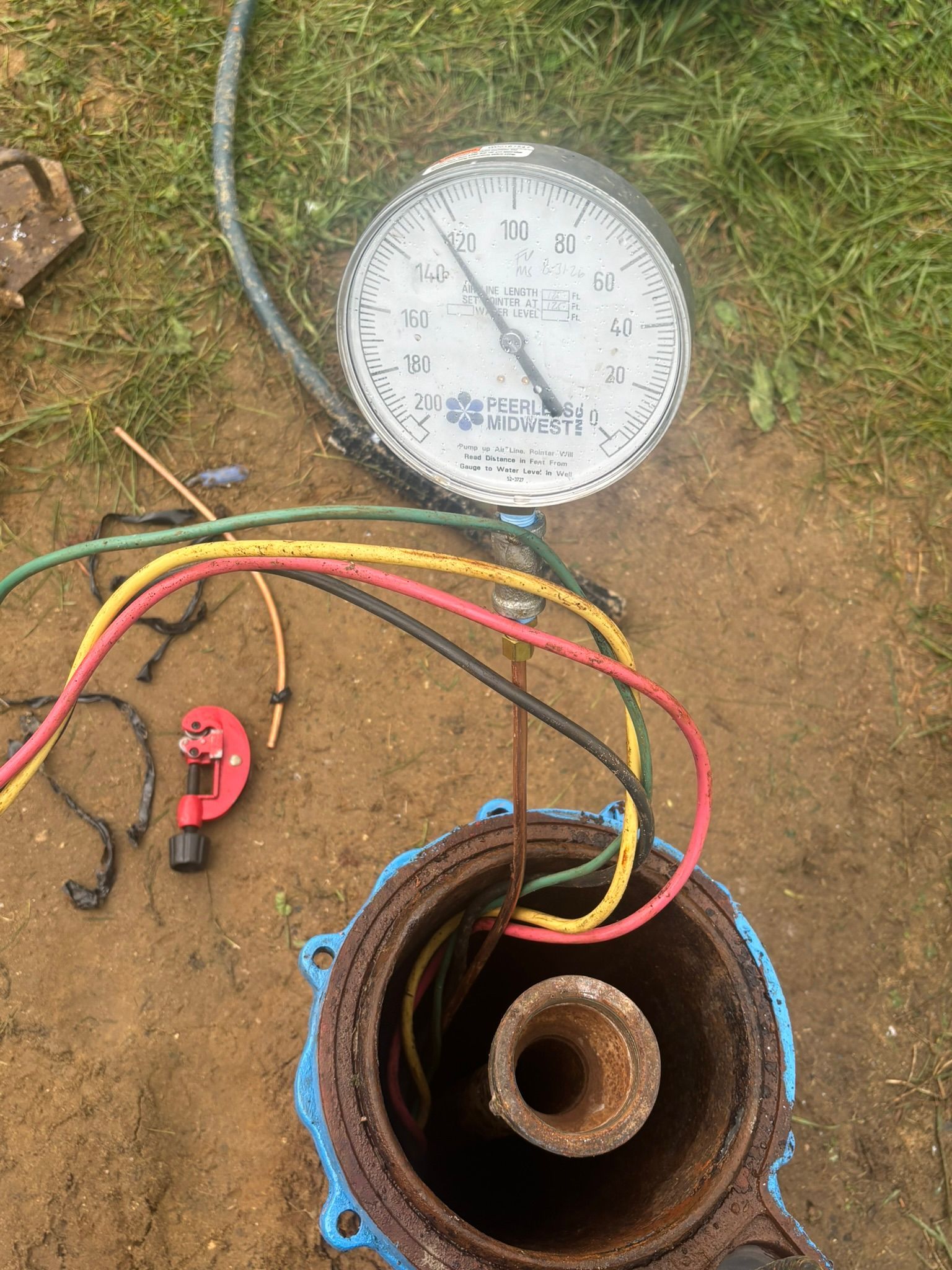 Pressure gauge with colored hoses attached to a rusted pipe opening on dirt ground.