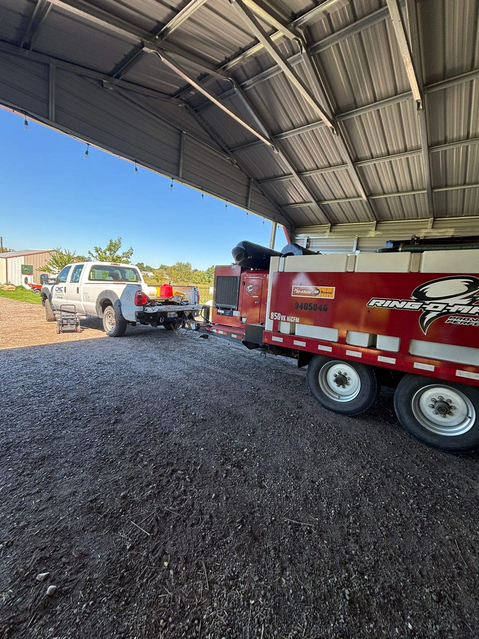 Dump trailer and pickup truck under a metal roof on a gravel lot