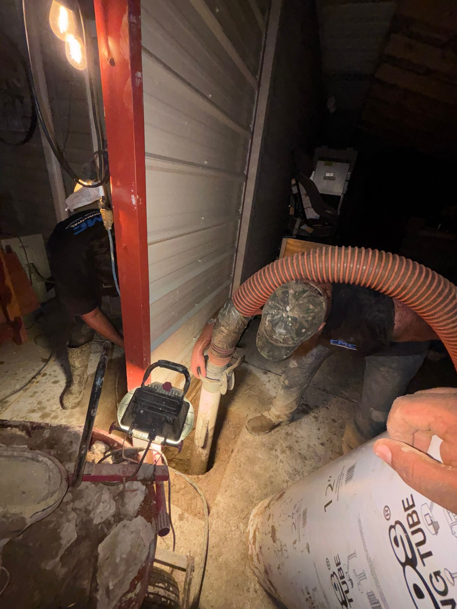Cluttered crawl space with red-framed door, orange hose, and dusty equipment on the floor.