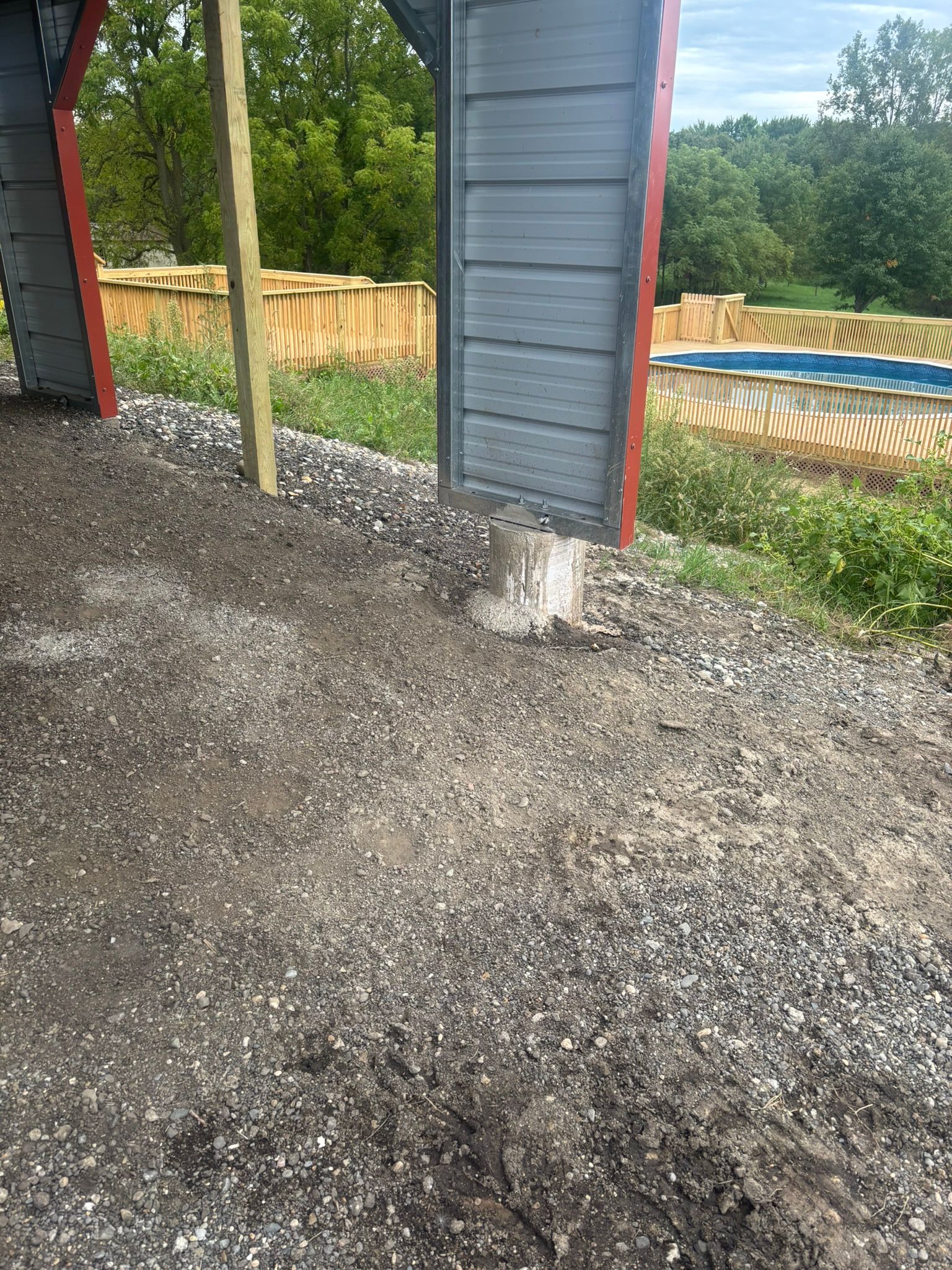 Gravel ground beside a gray building wall and red posts, with a fenced yard and trees in the background