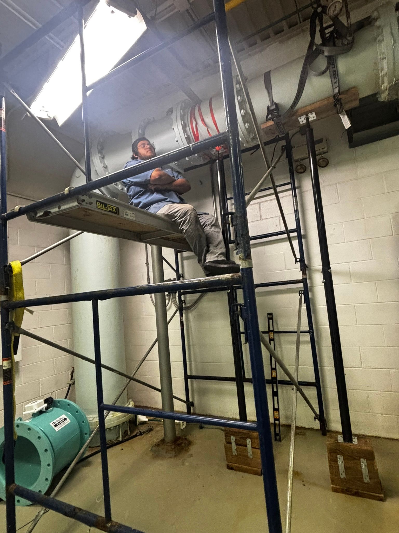 Worker on a black scaffold in an industrial room, with pipes, a green machine, and fluorescent lights above.