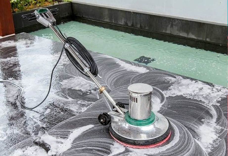 A floor buffer cleaning a dark stone patio next to a swimming pool, leaving white sudsy swirl patterns on the surface.