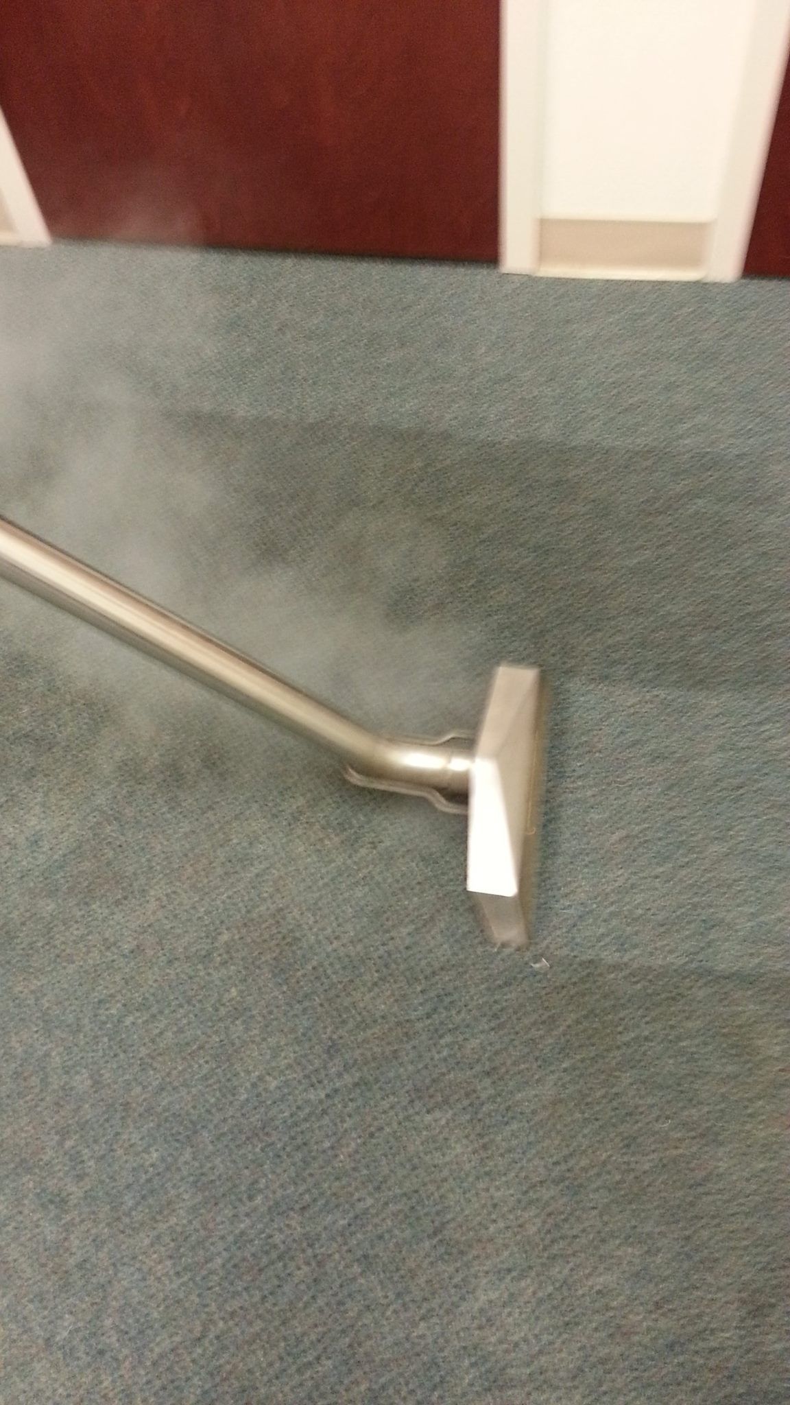 A metal carpet cleaning wand steaming a gray, textured carpeted surface.