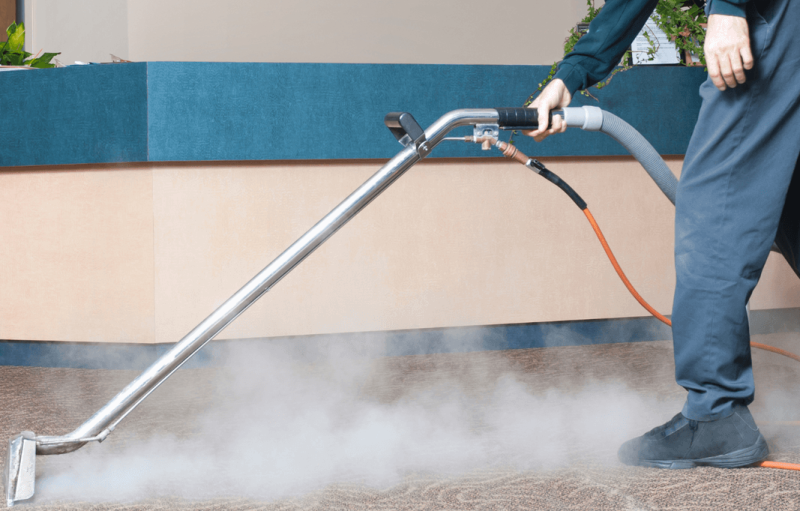 A person uses a steam cleaning wand to clean a carpet, releasing a cloud of vapor onto the floor.