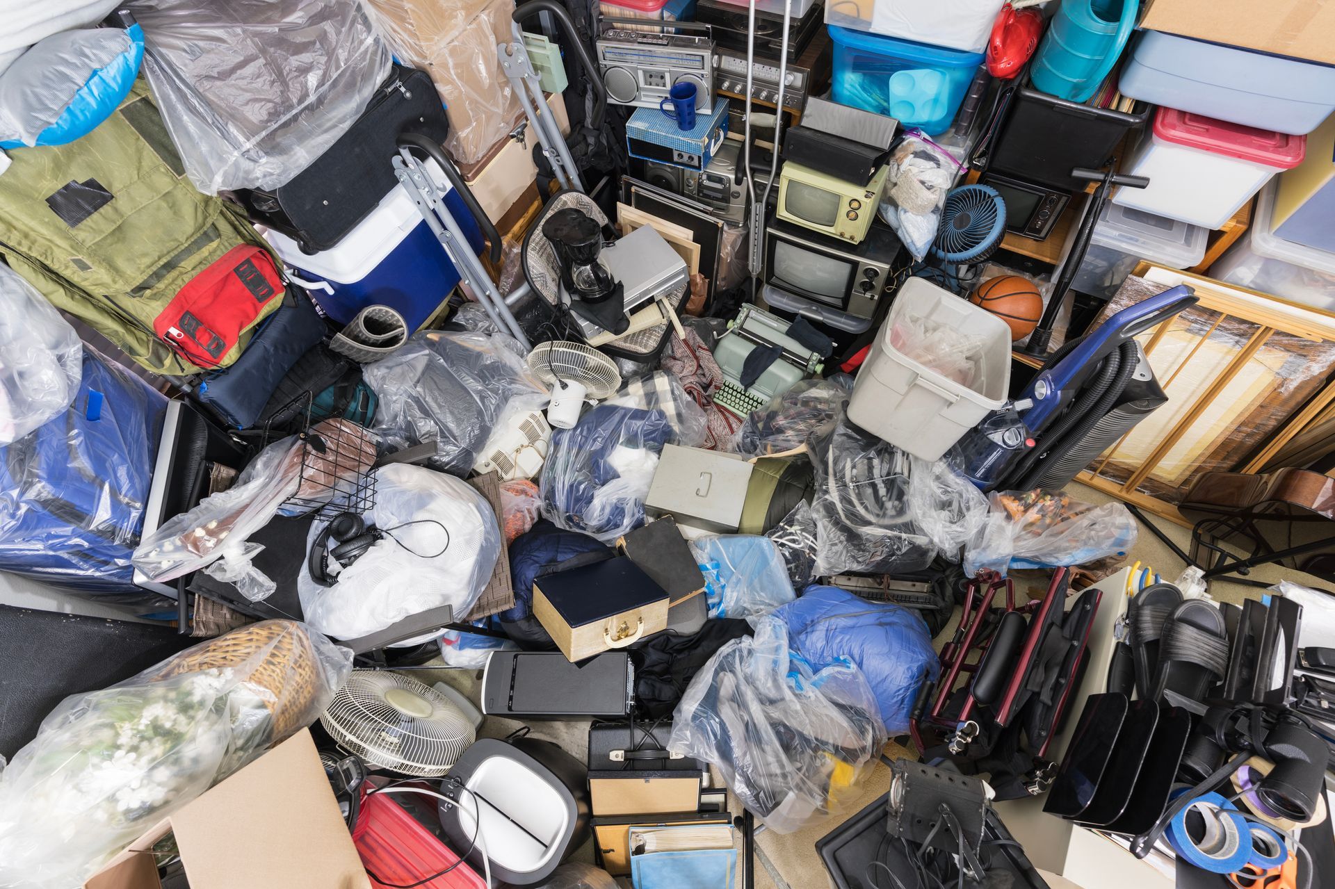 A storage unit packed floor to ceiling with a variety of miscellaneous items, boxes, electronics, and plastic-wrapped goods.