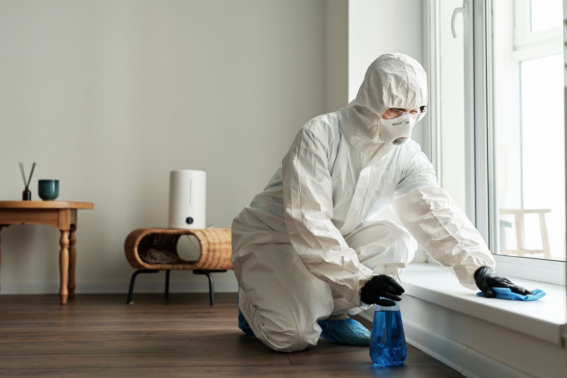 A person in a white hazmat suit and mask cleans a window ledge with a blue spray bottle and cloth in a minimalist room.