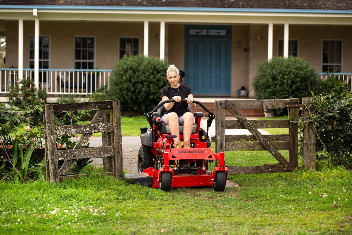 A Woman Is Riding a Lawn Mower in Front of A House — Cheshire Machinery in Bundaberg, QLD