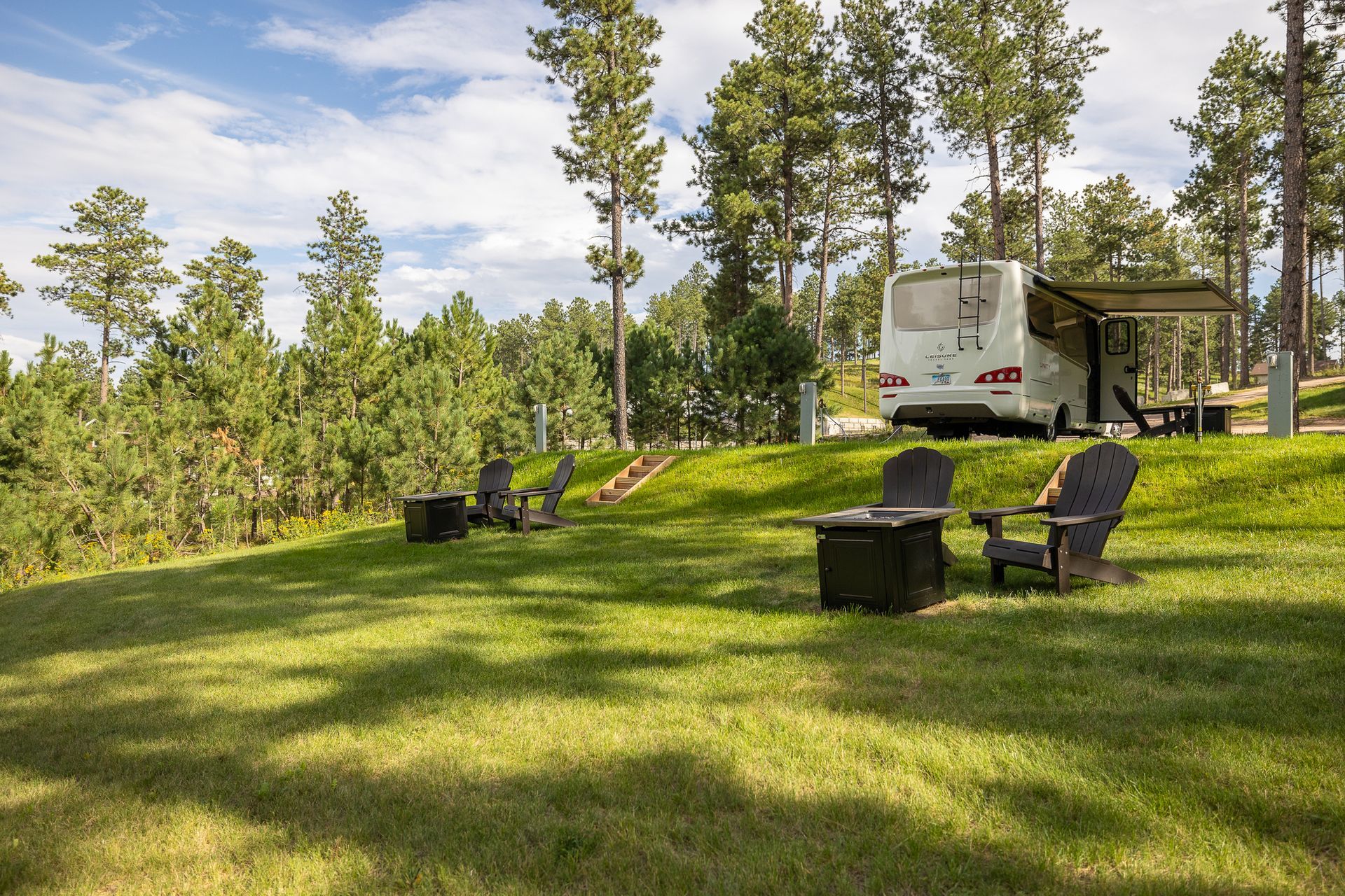 A rv is parked in a grassy field with chairs and a fire pit.