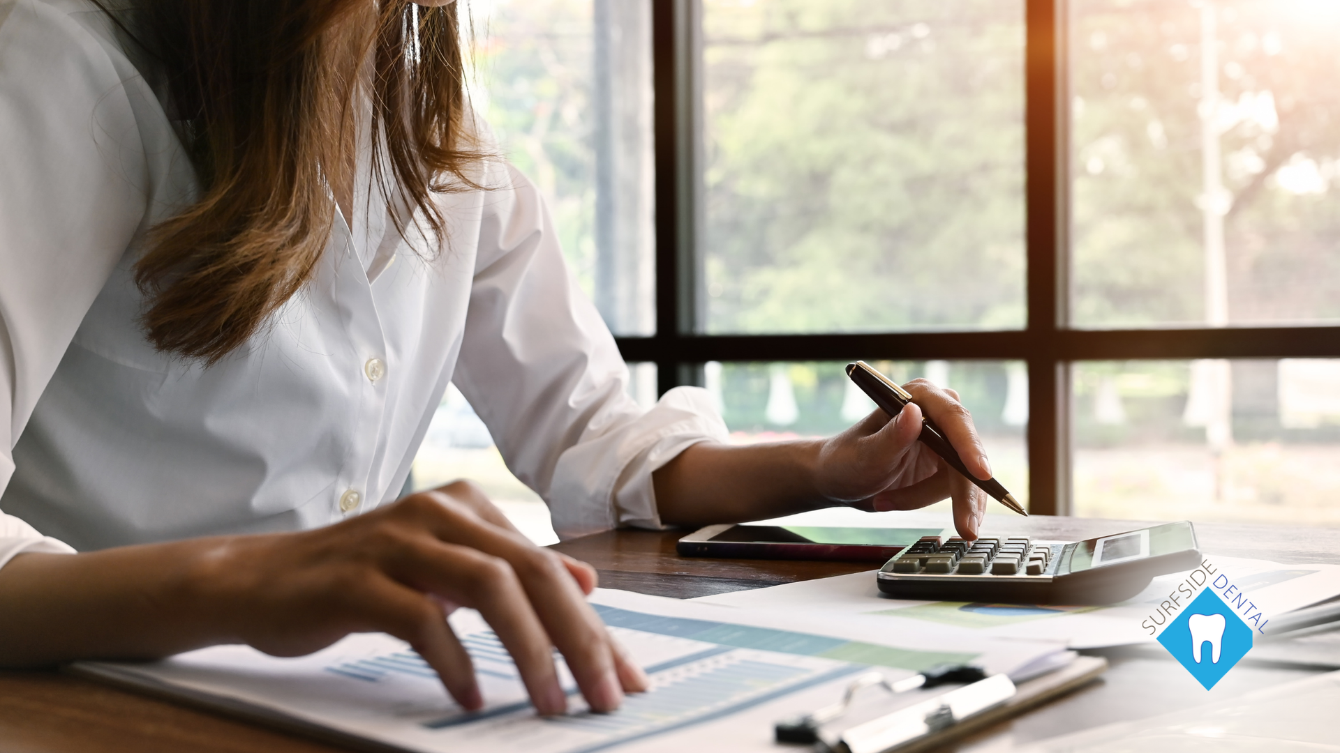 Woman using a calculator and reviewing documents at a desk near a window.