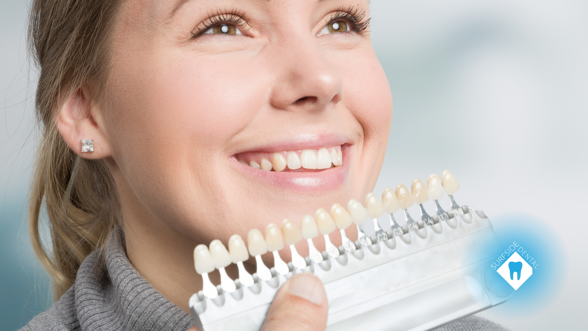 Woman comparing teeth to a shade guide for a dental procedure. She smiles in a medical setting.
