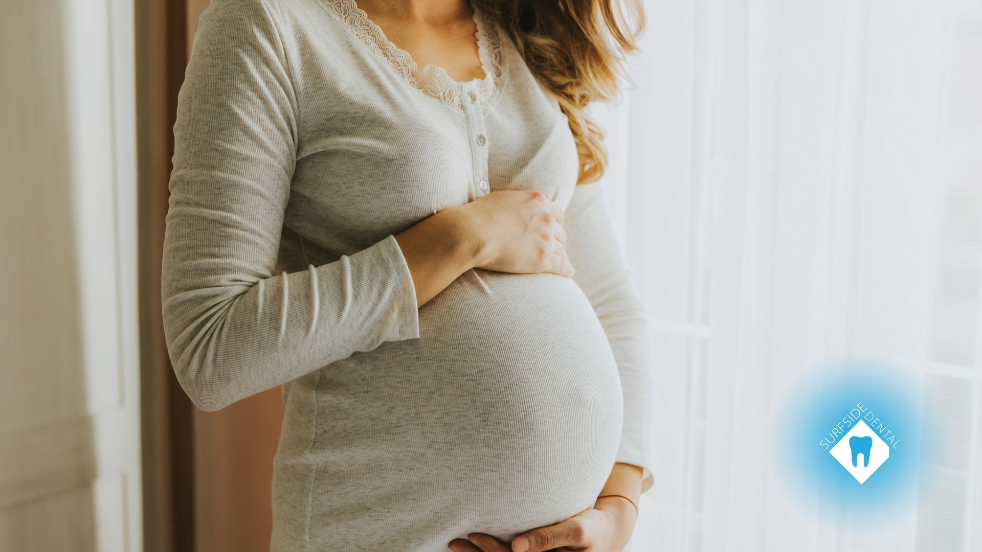 A pregnant woman is standing in front of a window holding her belly.