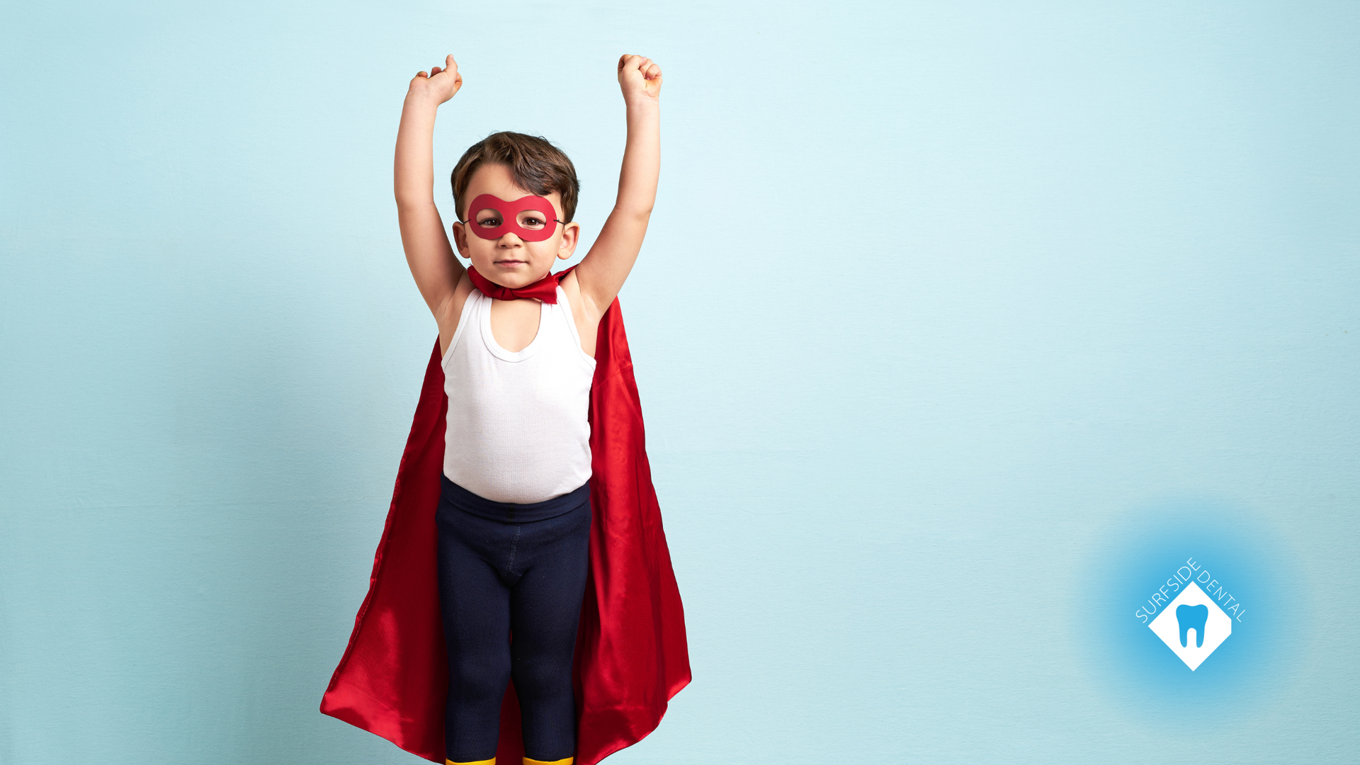 A young boy wearing a red cape and mask is standing with his arms in the air.