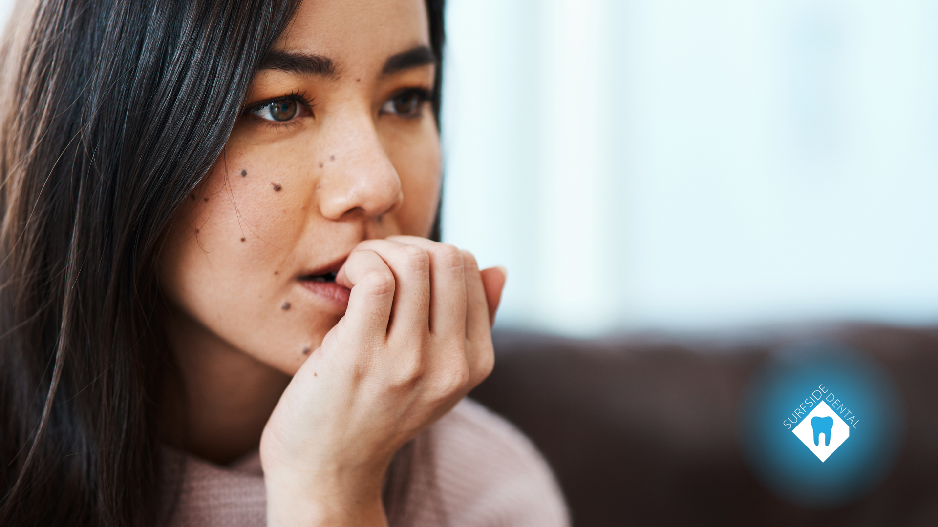 Woman biting nails, looking thoughtful; indoor setting, with a blue tooth icon.