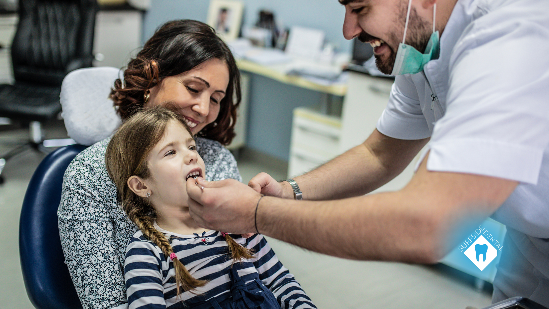 A little girl is sitting in a dental chair while her mother looks on.