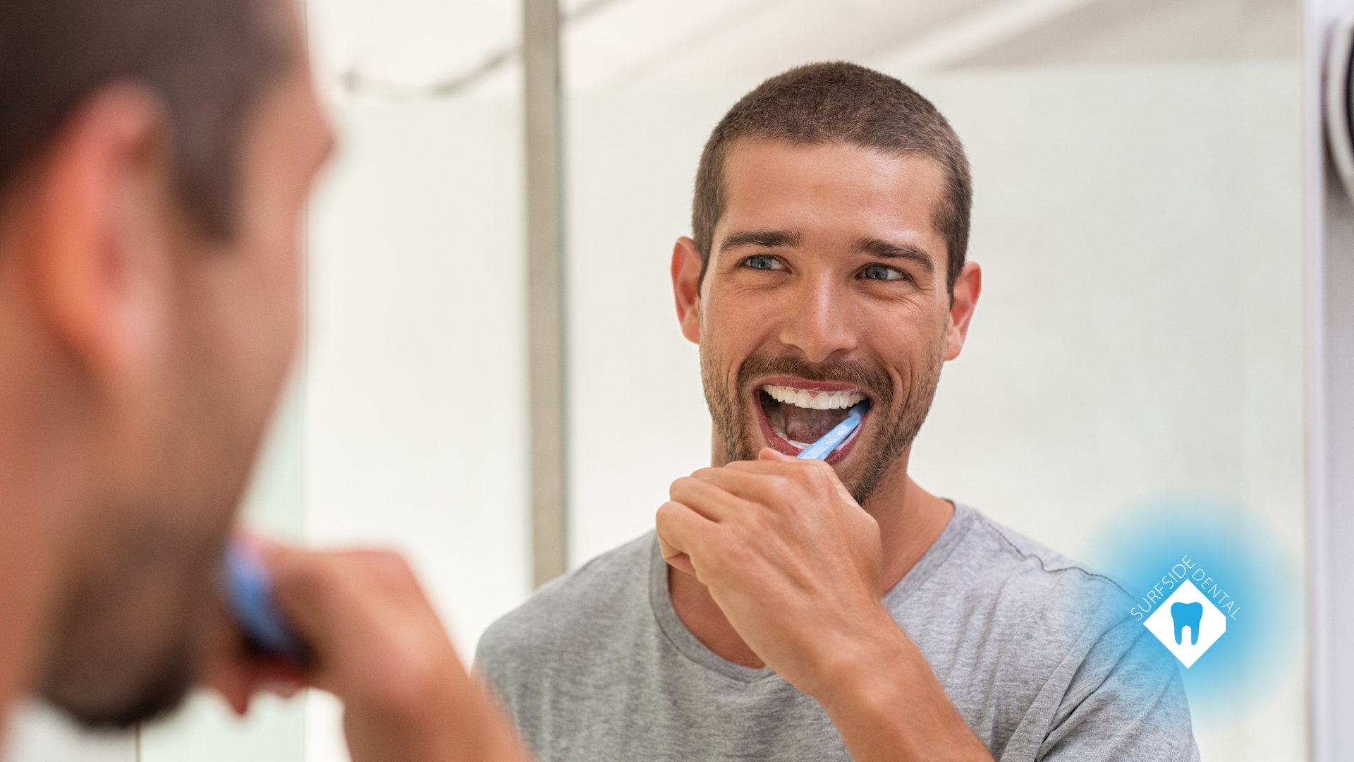 Man brushing teeth in the mirror; white teeth, open mouth, gray shirt, bathroom setting.
