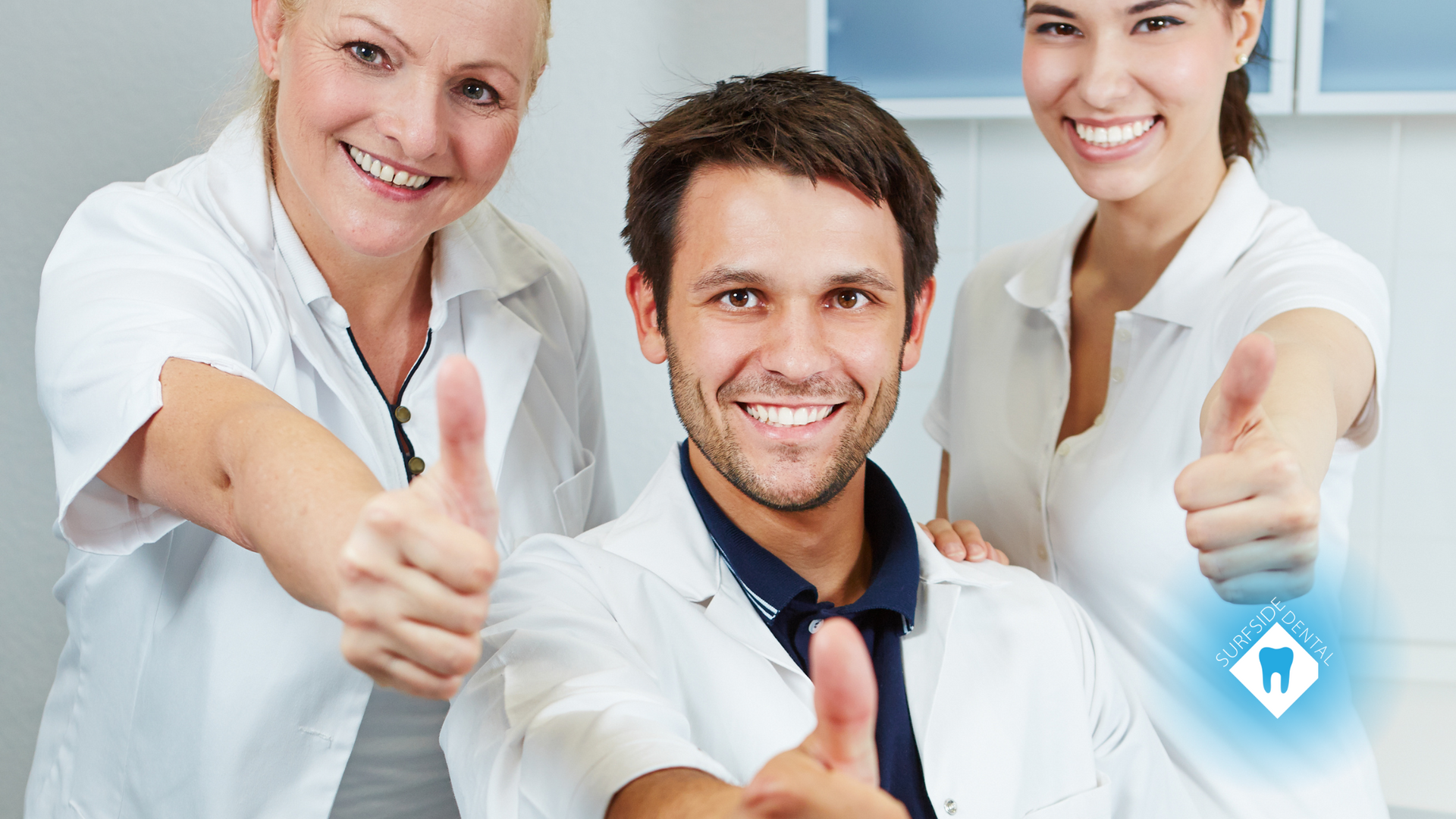Three smiling dental professionals give thumbs up, in a dental office setting.