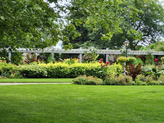 Lush green park lawn with flowering shrubs, a white trellis, and large trees in the background