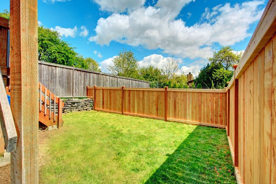 Sunny fenced backyard with green lawn, wooden deck stairs, and blue sky with clouds.