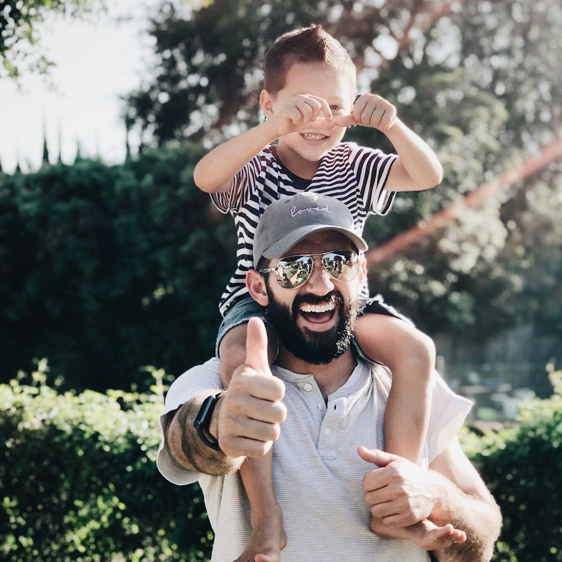 Man with a beard gives a thumbs up while carrying a child on his shoulders. Both are smiling outdoors.