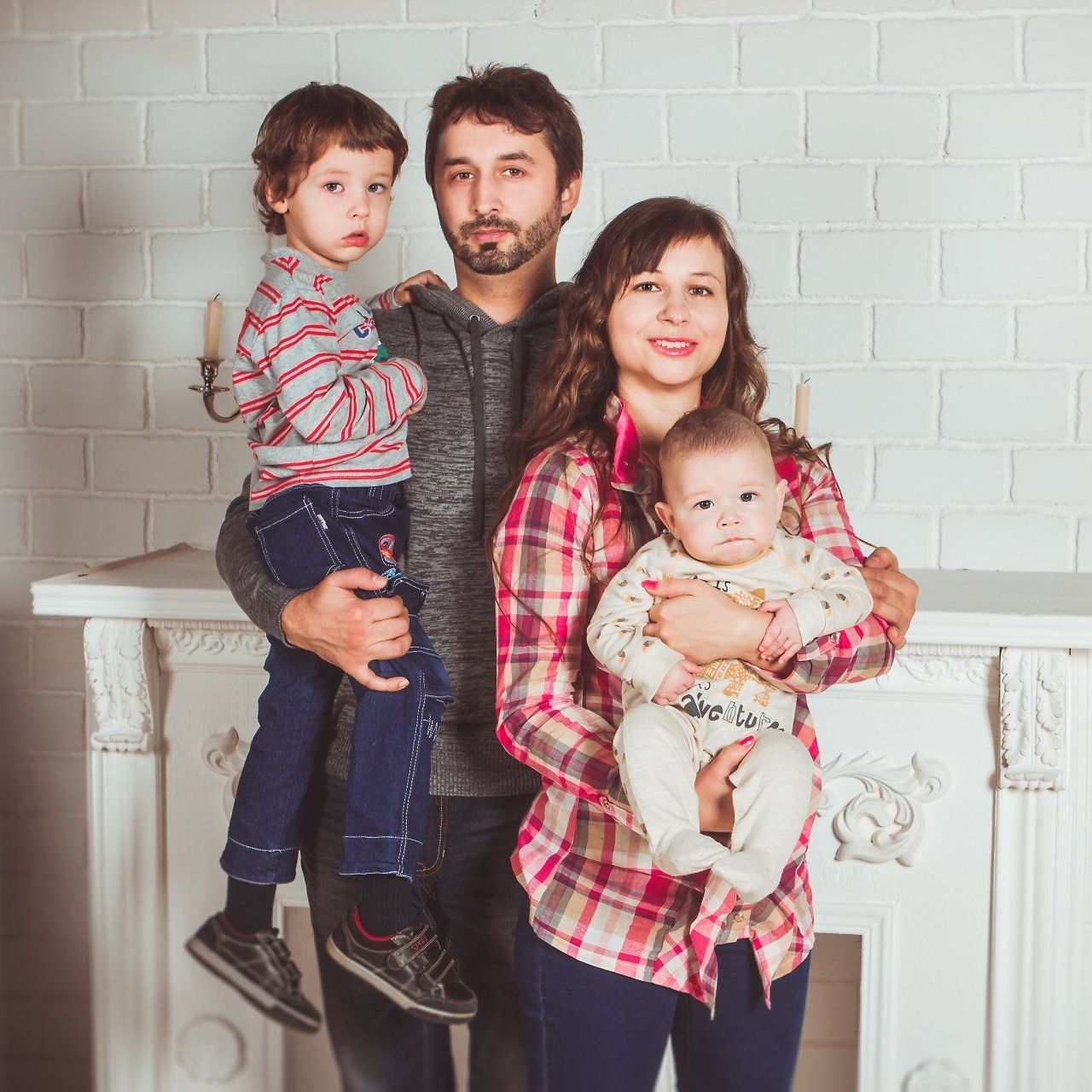 Family of four poses in front of a white brick wall; father holds a boy, mother holds a baby.
