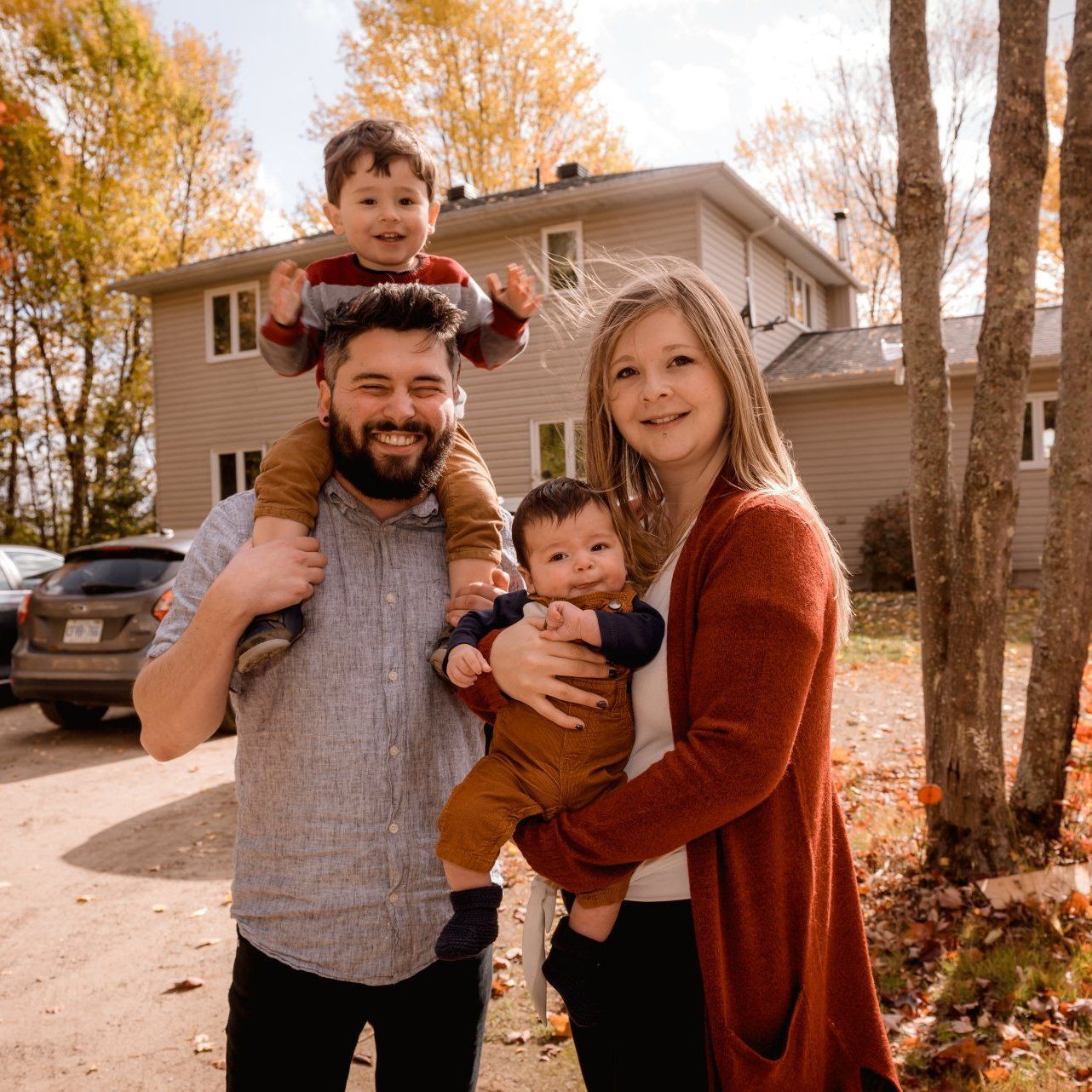 Family of four smiling in front of a house, father carrying a child, other child on his shoulders, fall colors.