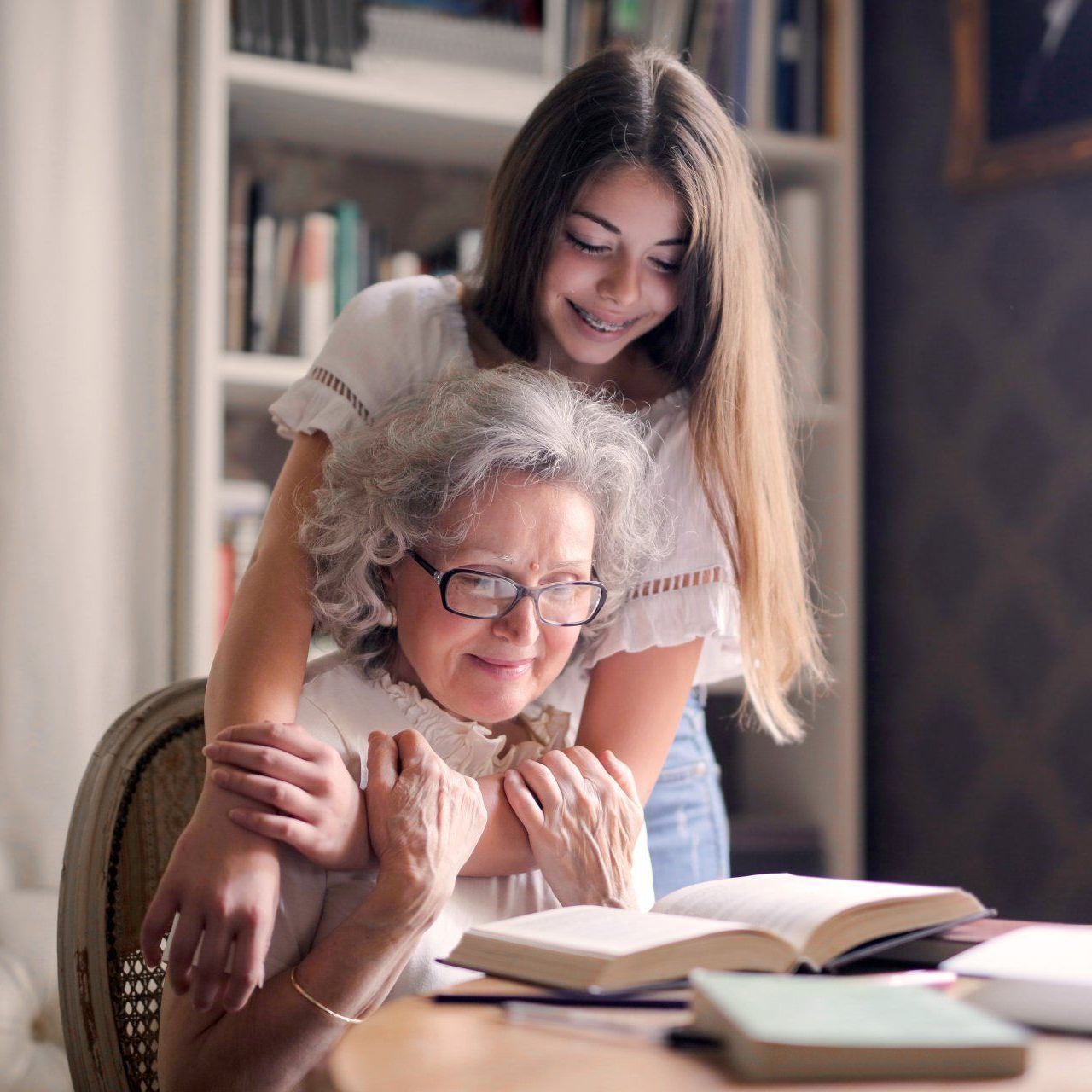 Teen girl hugs a smiling older woman seated at a table with books, in a library.